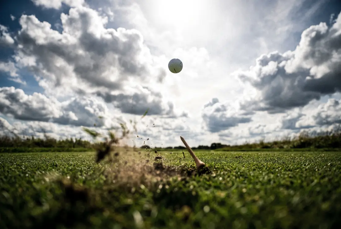 A golf ball climbing steeply into a cloudy sky, shot from low and behind with turf blurred in the foreground.