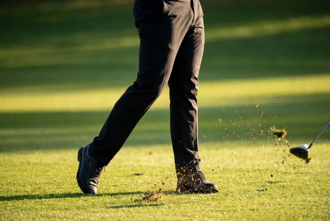 A golfer's lower body at impact, the back heel lifted clear of the ground and turf spraying forward.